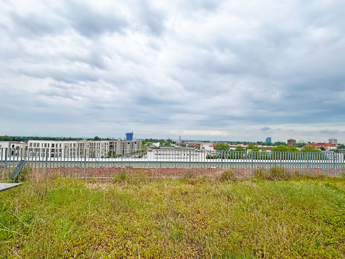 Cityscape view from a green rooftop with a metal fence under a cloudy sky.