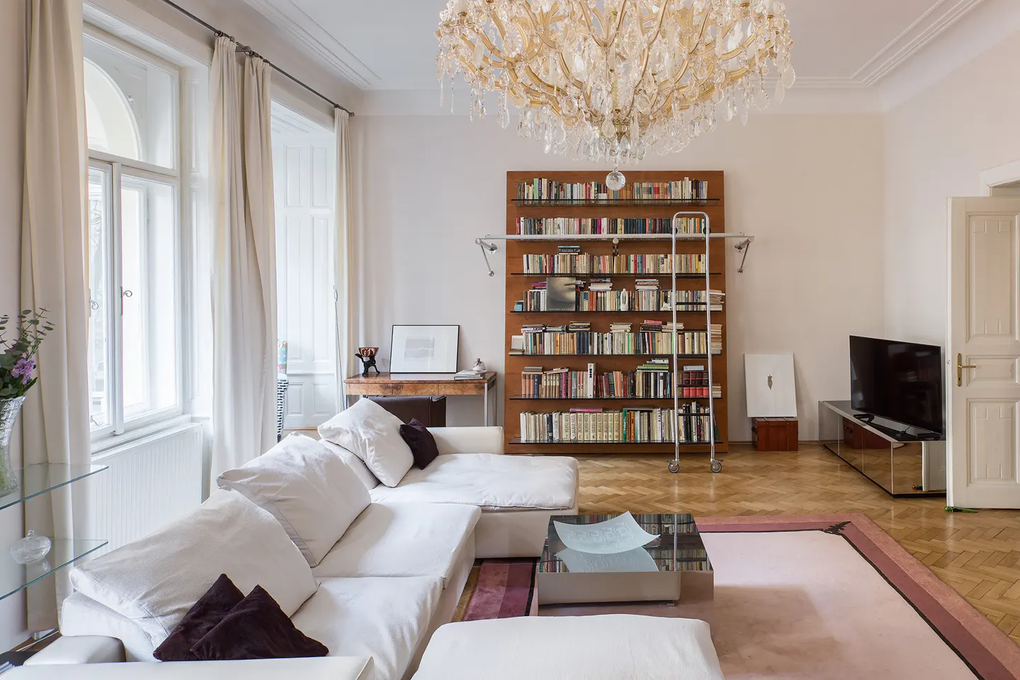 Bright living room with white walls, a large chandelier, and a bookshelf filled with books. A white sofa and a glass coffee table sit on a pink rug.