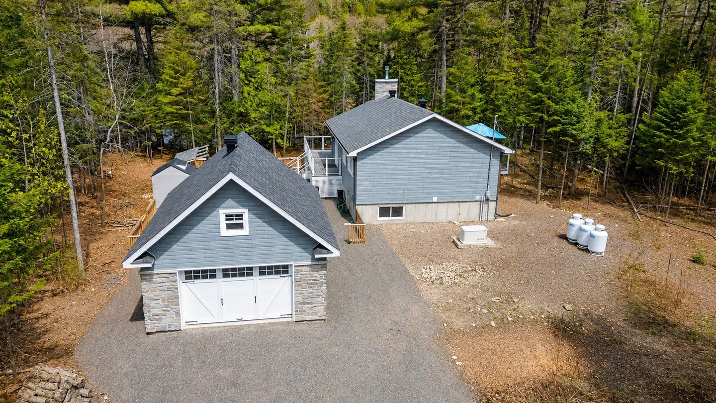 Aerial view of a light blue house and garage with white doors, surrounded by green trees. A gravel driveway leads to the buildings.
