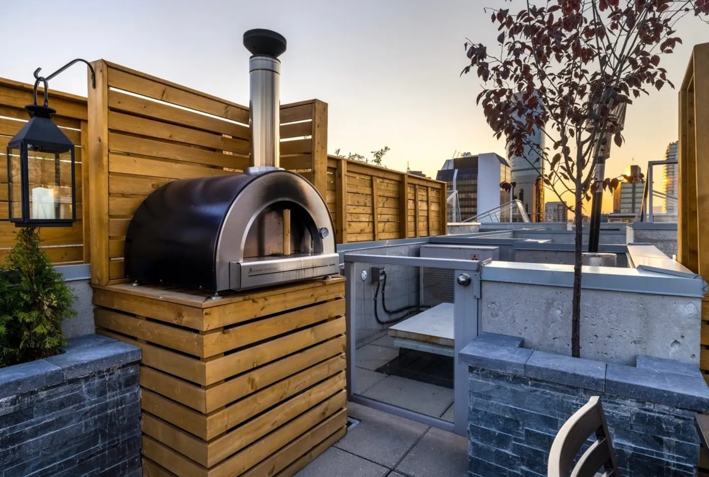 Rooftop patio with a black pizza oven on a wooden stand, a lantern, and a small tree. City buildings are visible in the background.