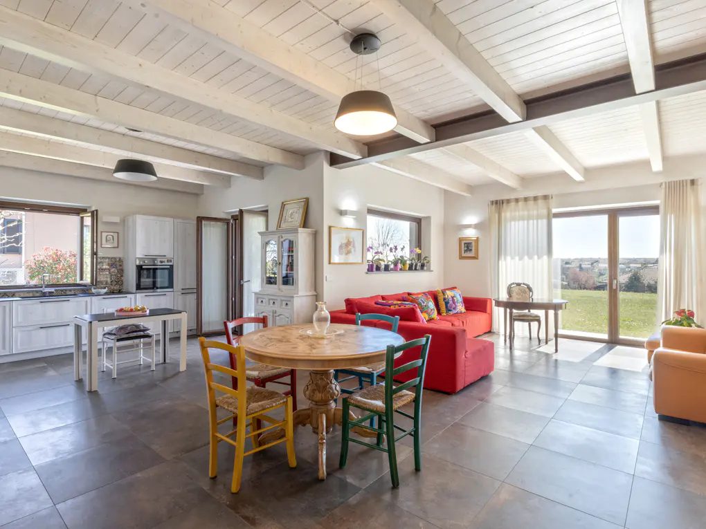Open-concept living space with a kitchen, dining table with colorful chairs, and a red sofa. Large windows overlook a green lawn.