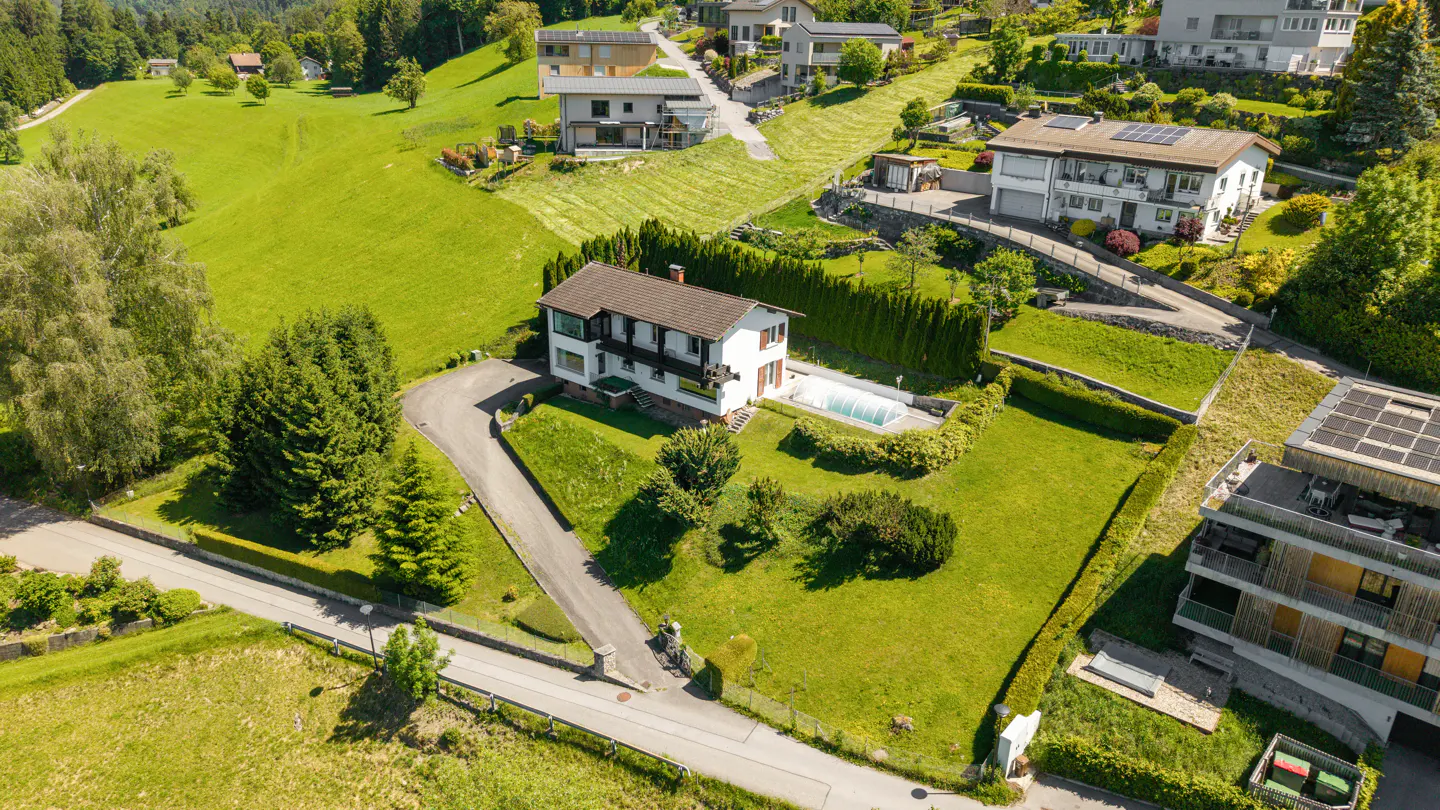 Aerial view of a white house with a pool, surrounded by green grass and trees on a hillside.