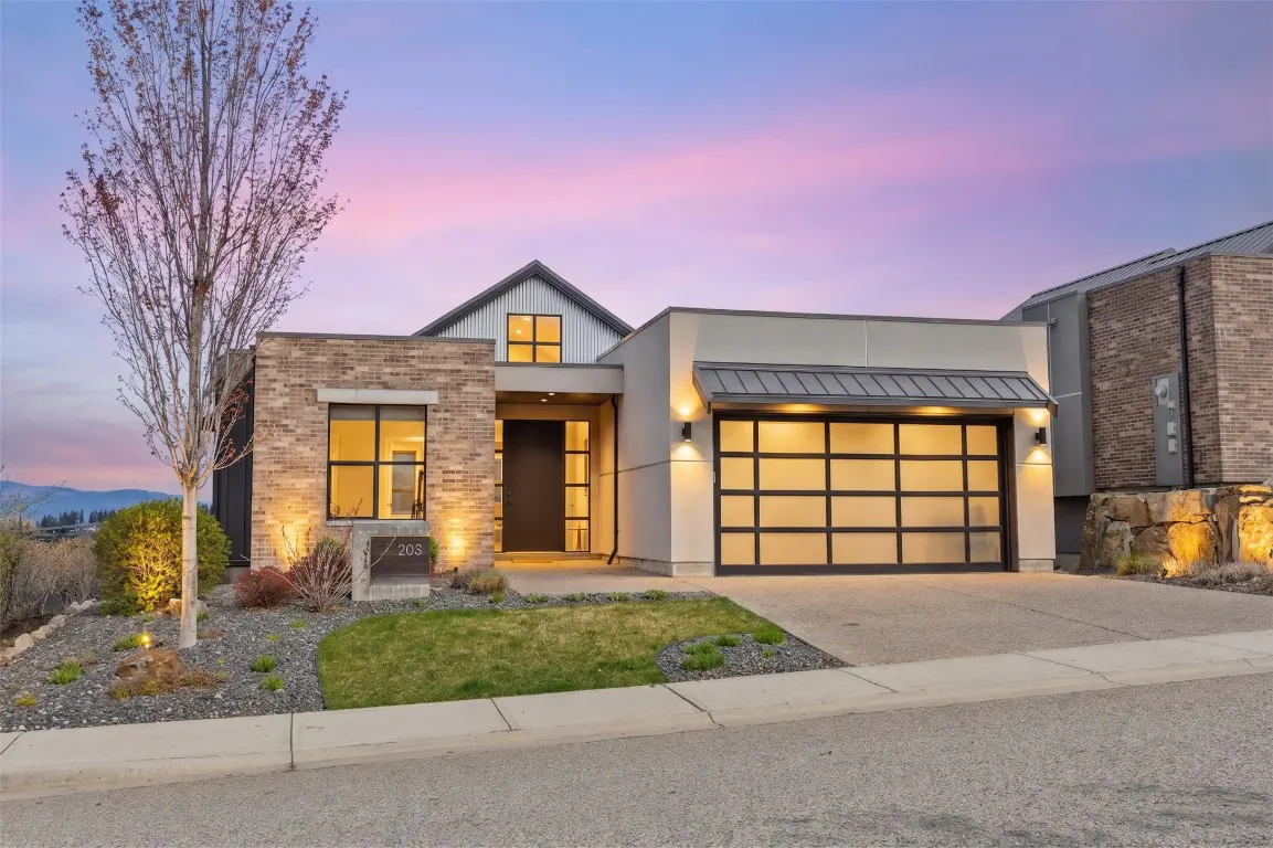 Modern home exterior at dusk with brick and gray siding, a glass garage door, and a tree in the front yard.