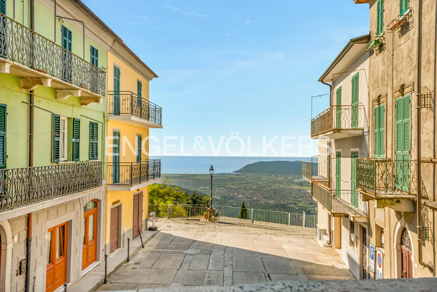 Street view of colorful buildings with balconies overlooking a valley and the sea under a blue sky.