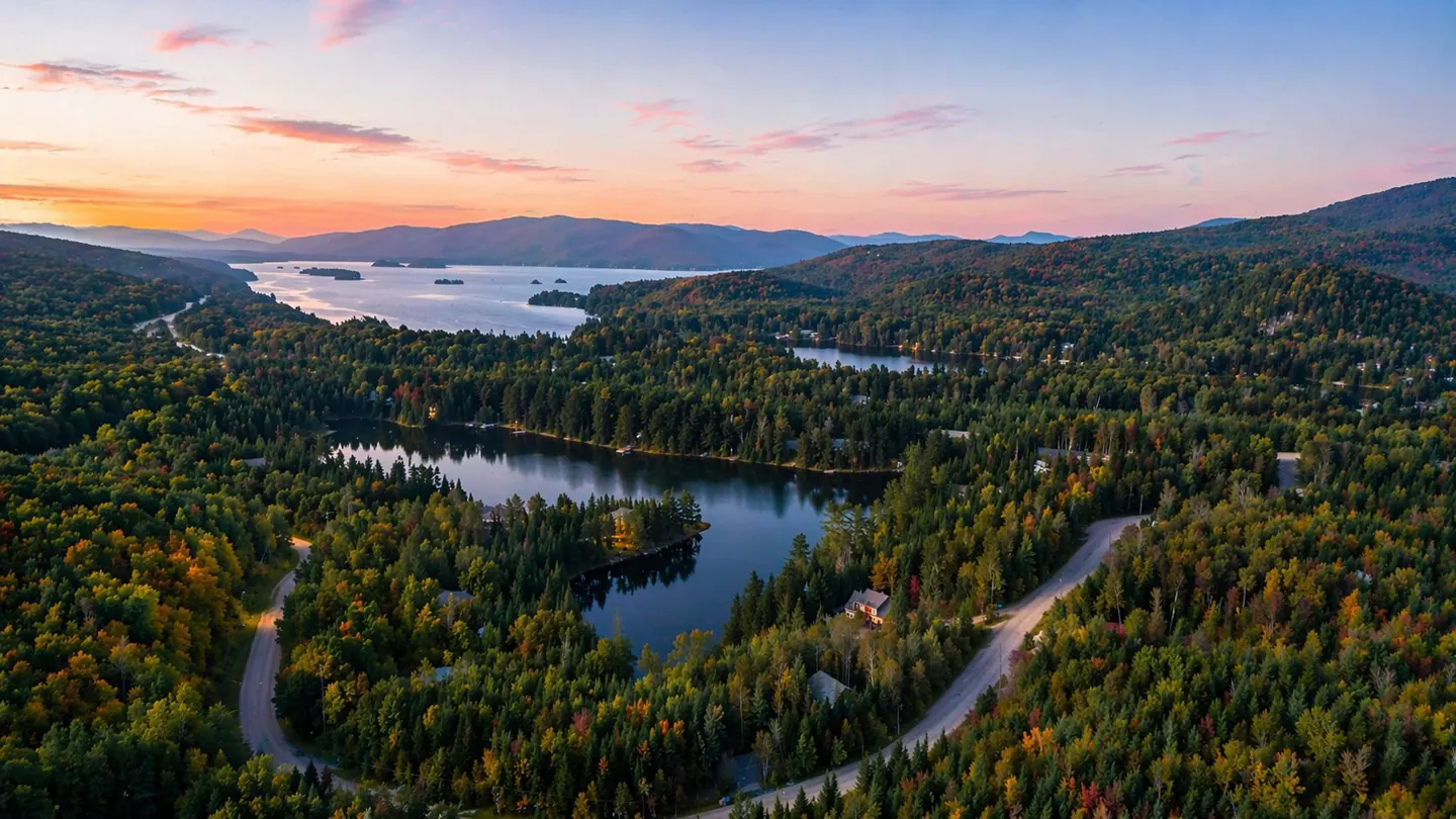 Aerial view of a lake surrounded by green forests and mountains at sunset. A winding road cuts through the trees.