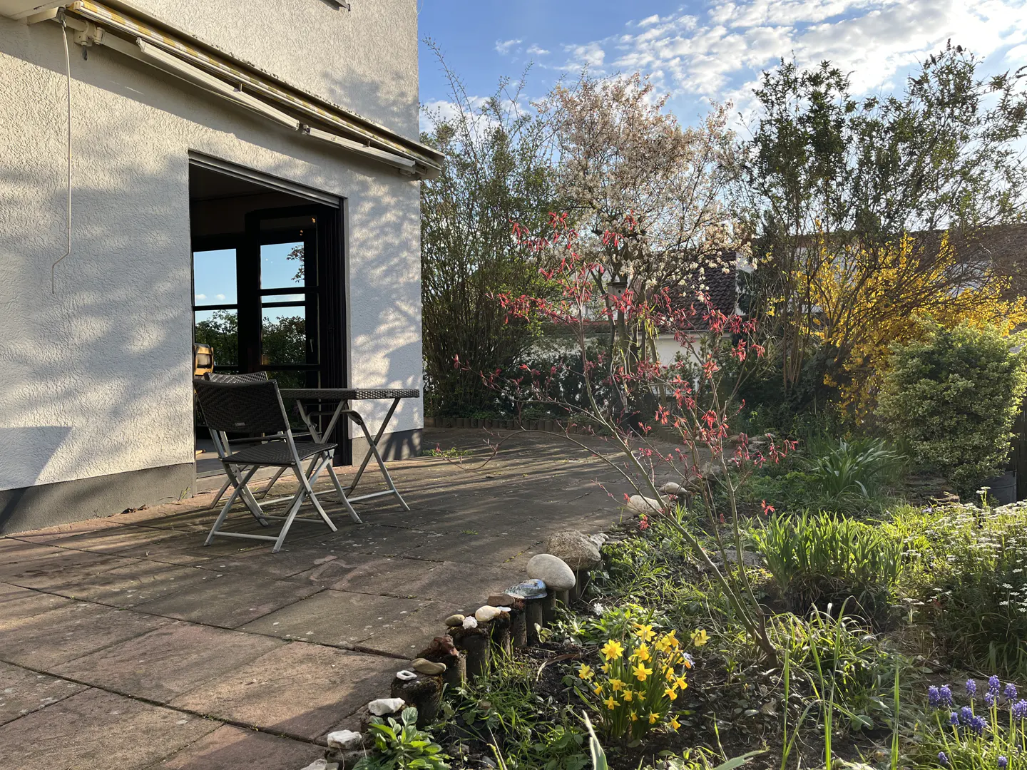 A patio with a table and chairs sits outside a white house, with a garden full of colorful flowers in the foreground.