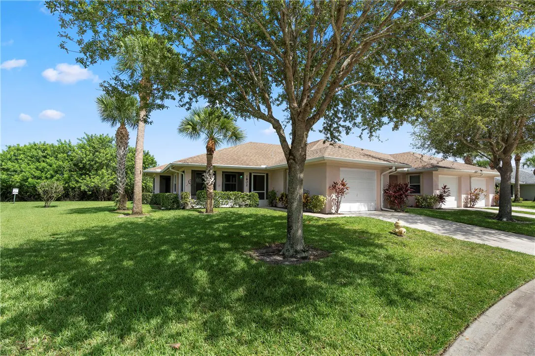 Single-story beige house with a tan roof, palm trees, and a green lawn under a clear blue sky.
