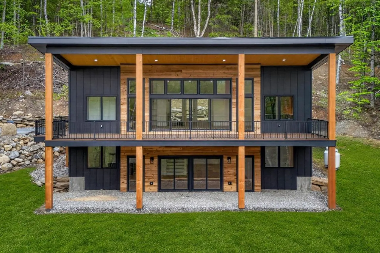 Two-story modern house with black siding, wood accents, and large windows. The house has a deck and is surrounded by green grass and trees.