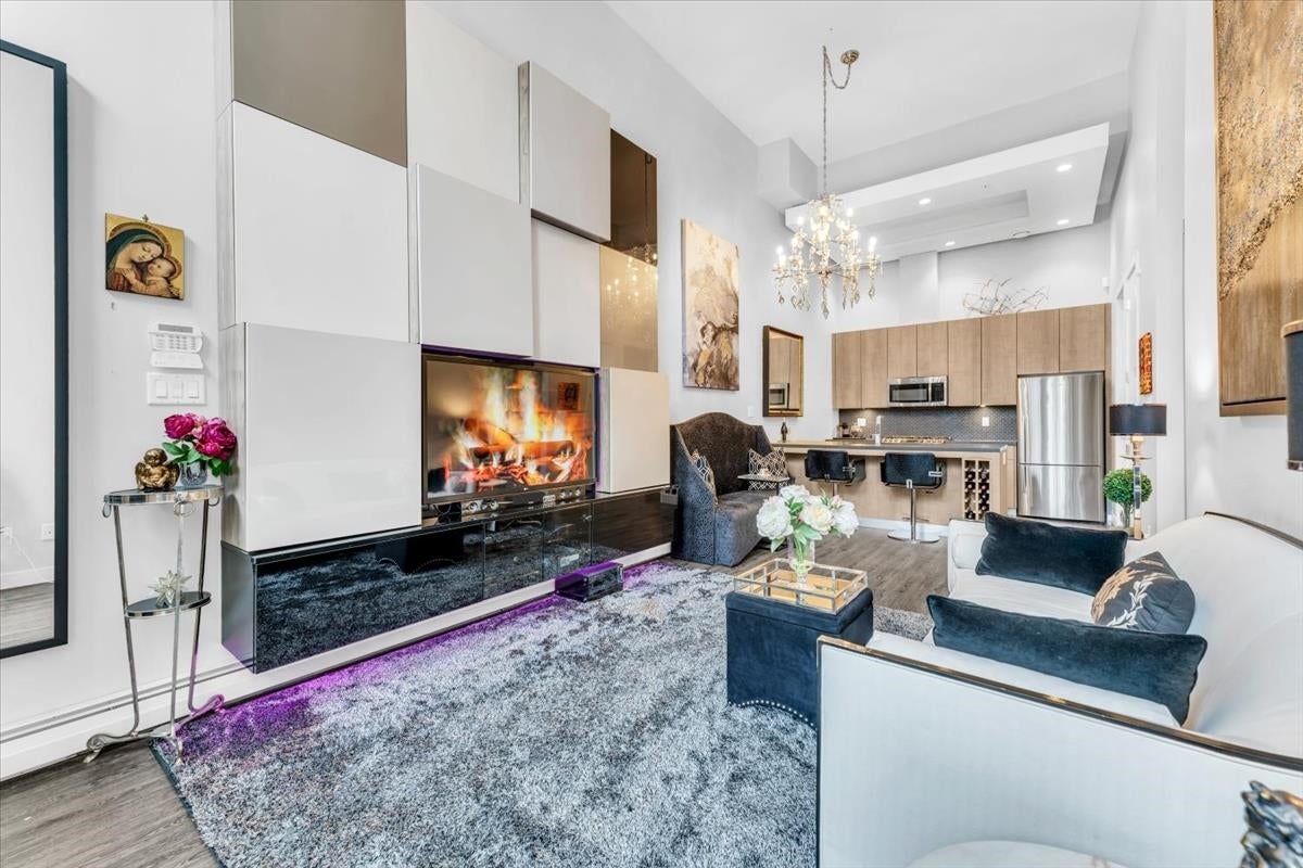 Modern living room with a fireplace wall, plush grey rug, white sofa, and an open kitchen with a crystal chandelier.