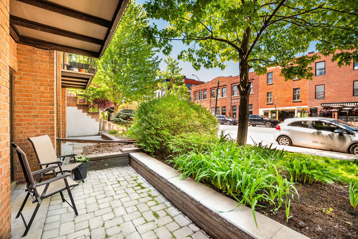 Brick building with a patio featuring two chairs, a brick walkway, and green plants. A street with cars and buildings is visible in the background.