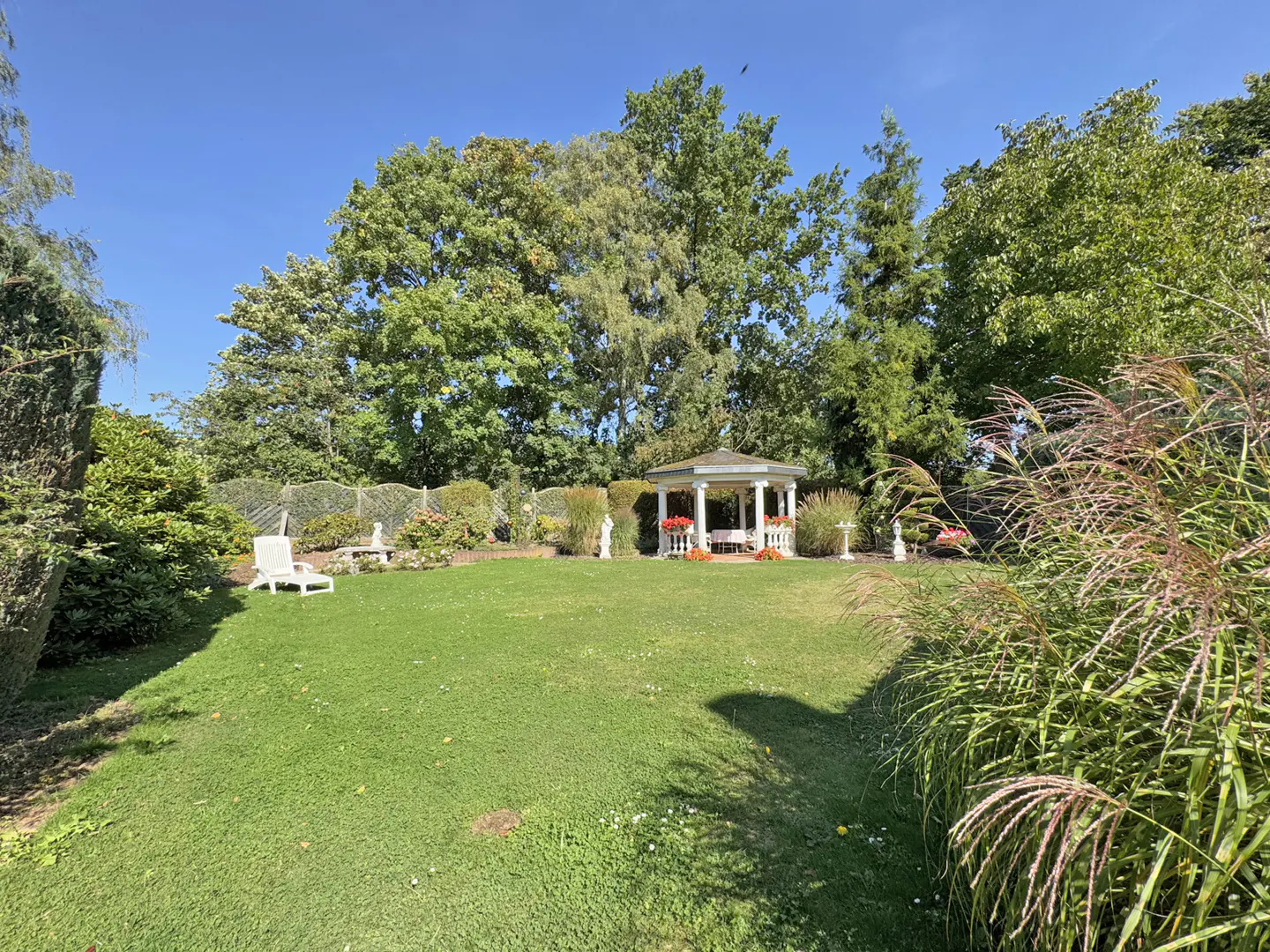 Lush green lawn with a white gazebo in the center, surrounded by trees and manicured bushes under a clear blue sky.