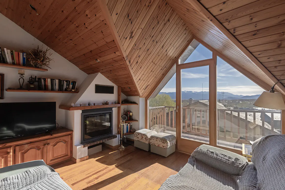 Attic living room with wood ceiling, fireplace, TV, and a view of mountains through a large window.