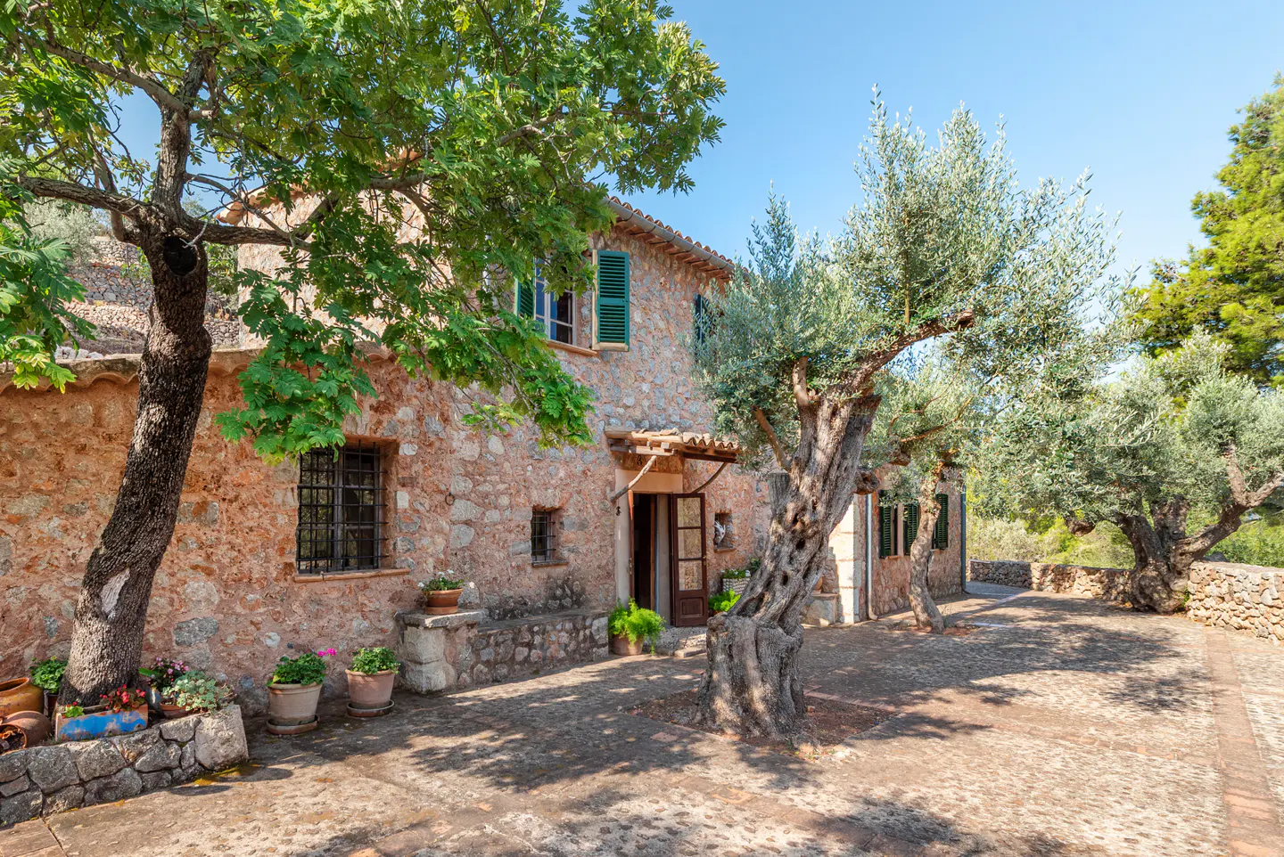 Stone house with green shutters, framed by trees. The courtyard is paved with stones. Blue sky.