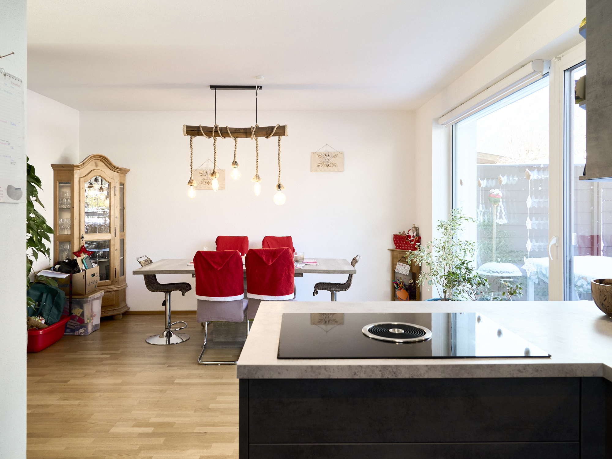 A bright, modern kitchen and dining area with wood floors. A dining table with chairs covered in red Santa hats sits under a rustic light fixture. A large window provides natural light.