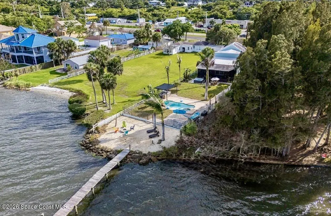 Aerial view of a waterfront property with a dock, pool, and sandy beach area. Lush green lawn and trees surround the house.