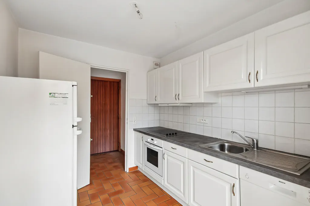 Bright kitchen with white cabinets, gray countertop, and stainless steel sink. A white refrigerator stands next to a doorway with a wooden door. The floor is tiled in a herringbone pattern.