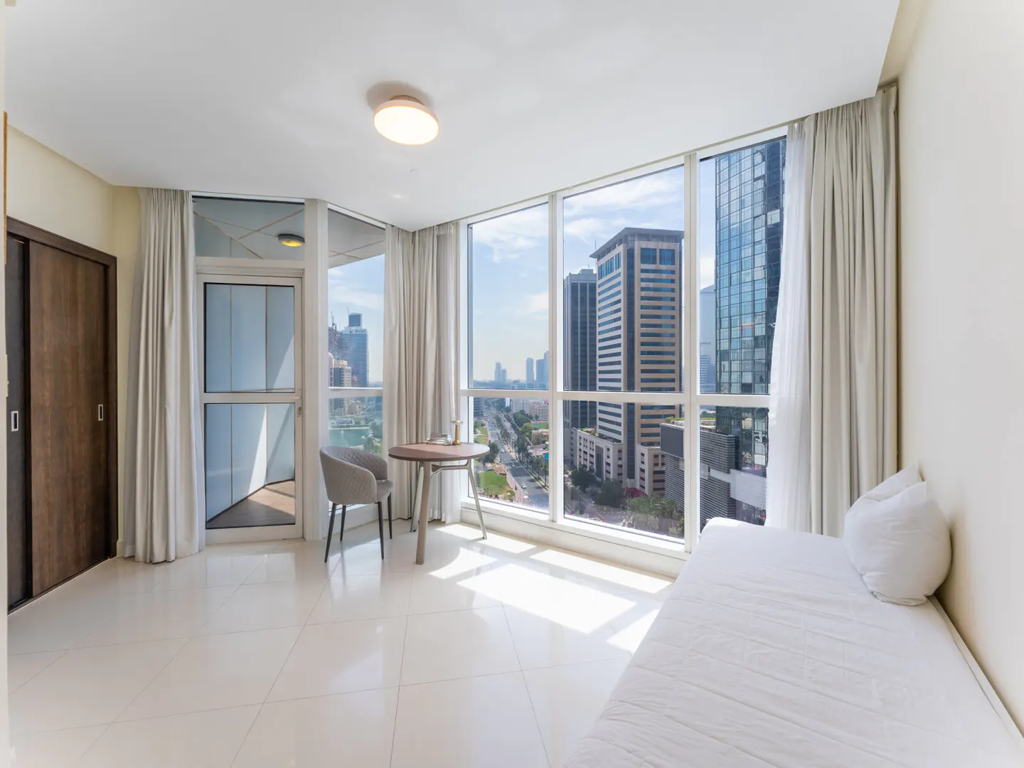 Bright apartment interior with a white daybed, table, chair, and floor-to-ceiling windows overlooking a city skyline.