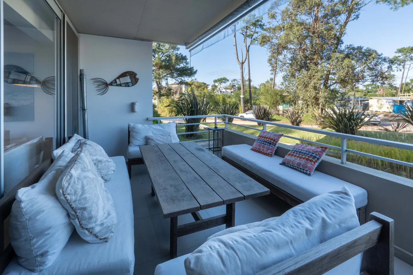 A bright balcony with a wooden table, white cushioned benches, and colorful pillows. A metal fish decoration hangs on the wall. Trees are visible in the background.