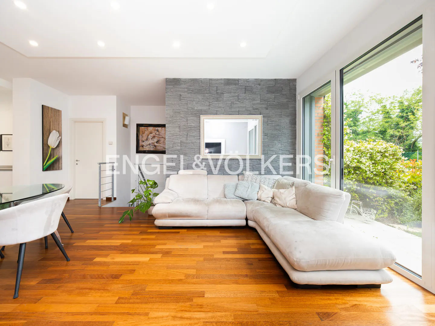 Bright living room with hardwood floors, a white sectional sofa, and a large window overlooking a green garden.