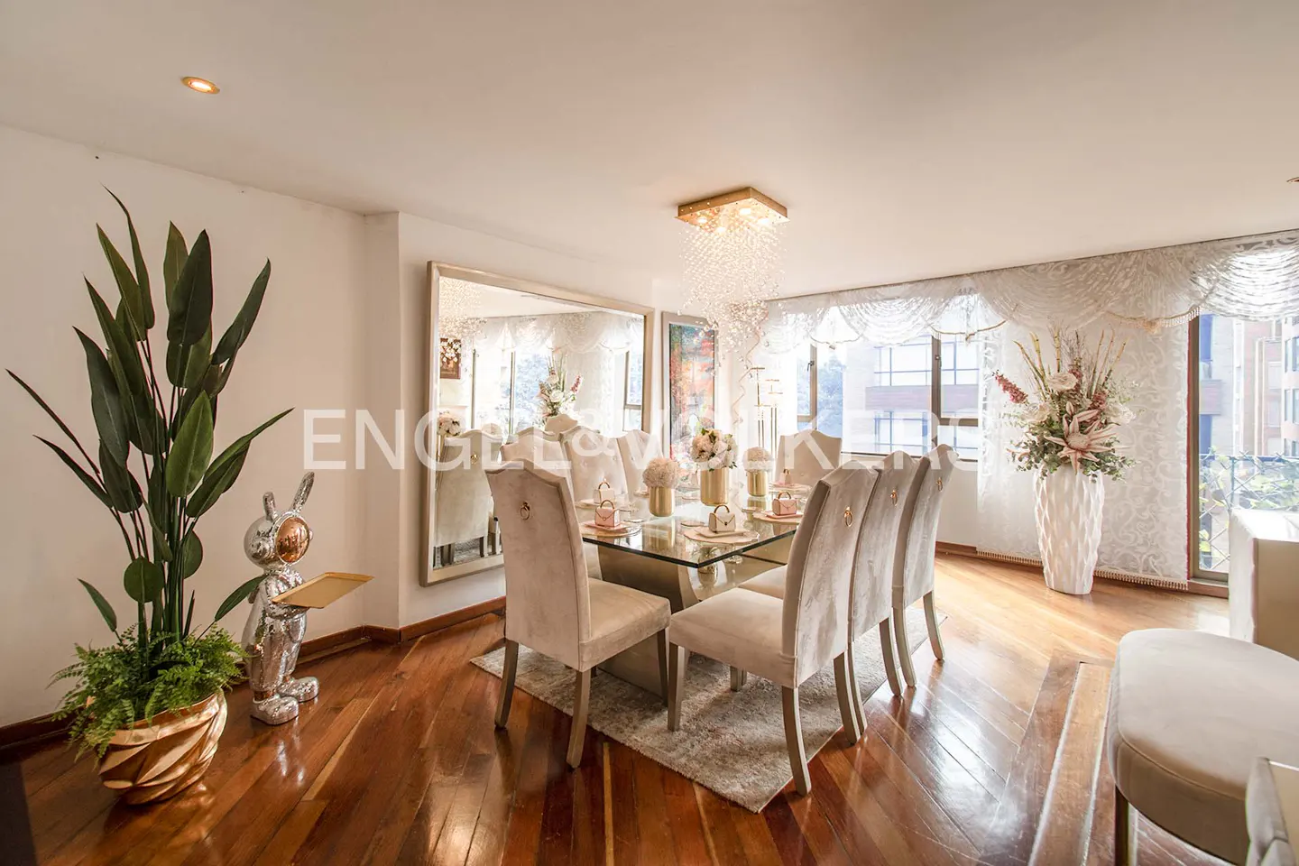 Bright dining room with wood floors, a glass table with six chairs, and a large window with sheer curtains. A large plant and silver rabbit statue are on the left.