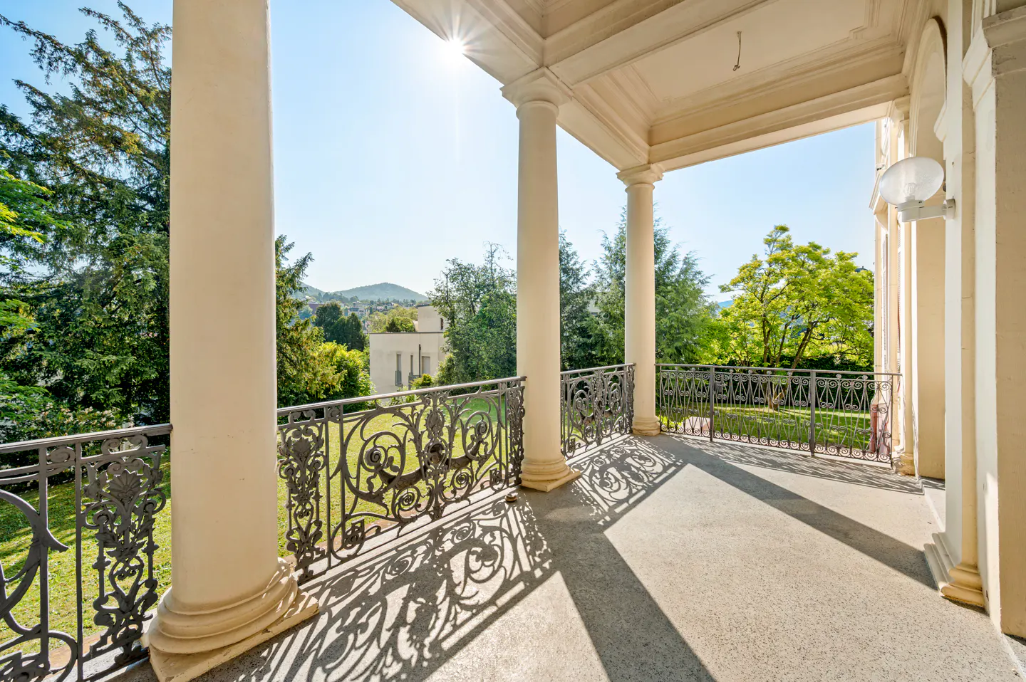 View from a covered porch with white columns and decorative iron railing overlooking a green lawn and trees on a sunny day.