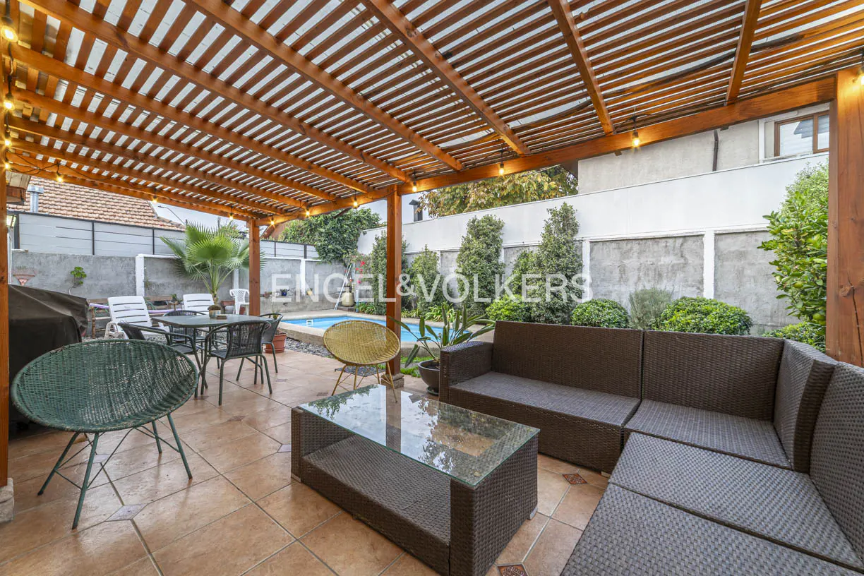 Outdoor patio with a wooden pergola, wicker sofa, glass coffee table, and chairs overlooking a backyard swimming pool.