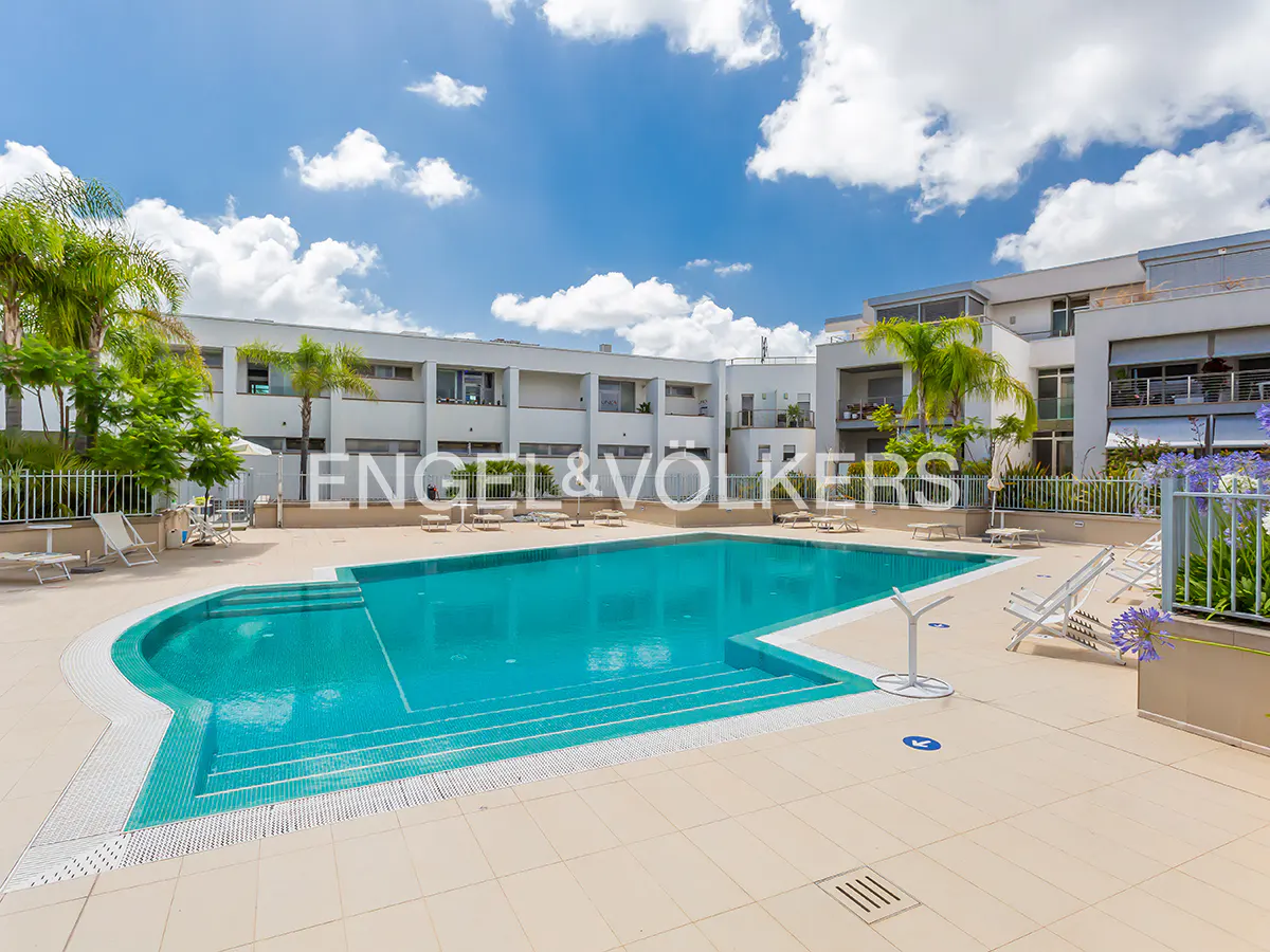 A turquoise swimming pool with steps, surrounded by a beige tiled deck, palm trees, and a modern apartment building.