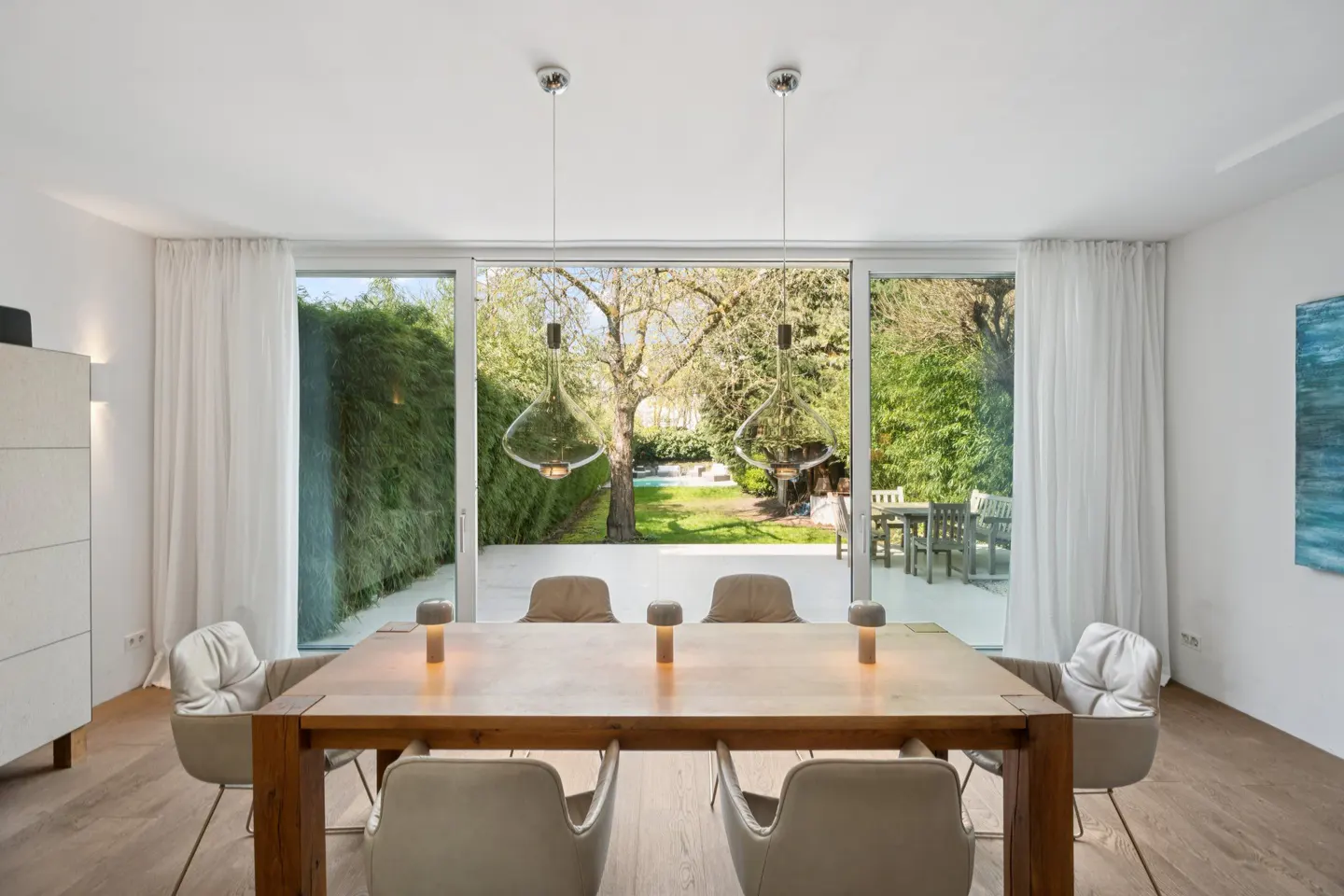 Bright dining room with a wooden table, six chairs, and three small lamps. A large glass door shows a green backyard with a table and chairs.