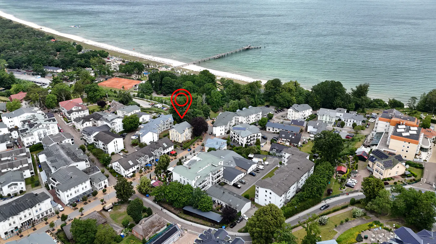 Aerial view of a coastal town with a red location pin marking a specific house near a sandy beach and a long pier.