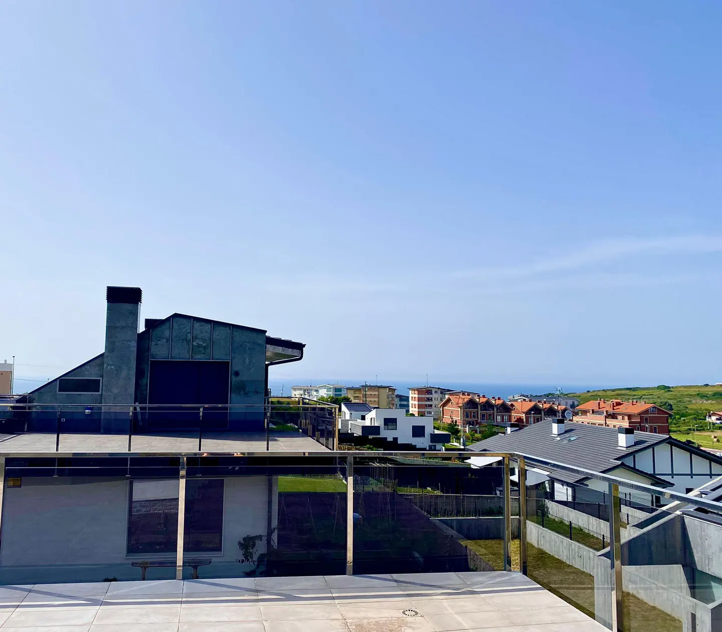 View from a rooftop terrace with glass railings, overlooking houses and the ocean under a clear blue sky.
