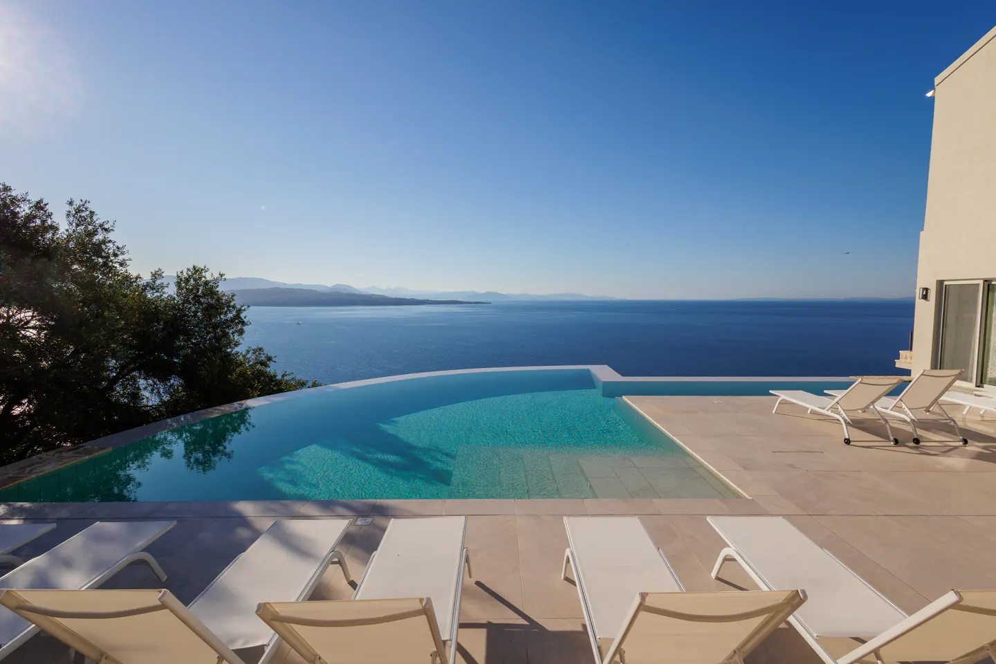 Infinity pool overlooking the ocean, with lounge chairs on the patio. Blue sky.