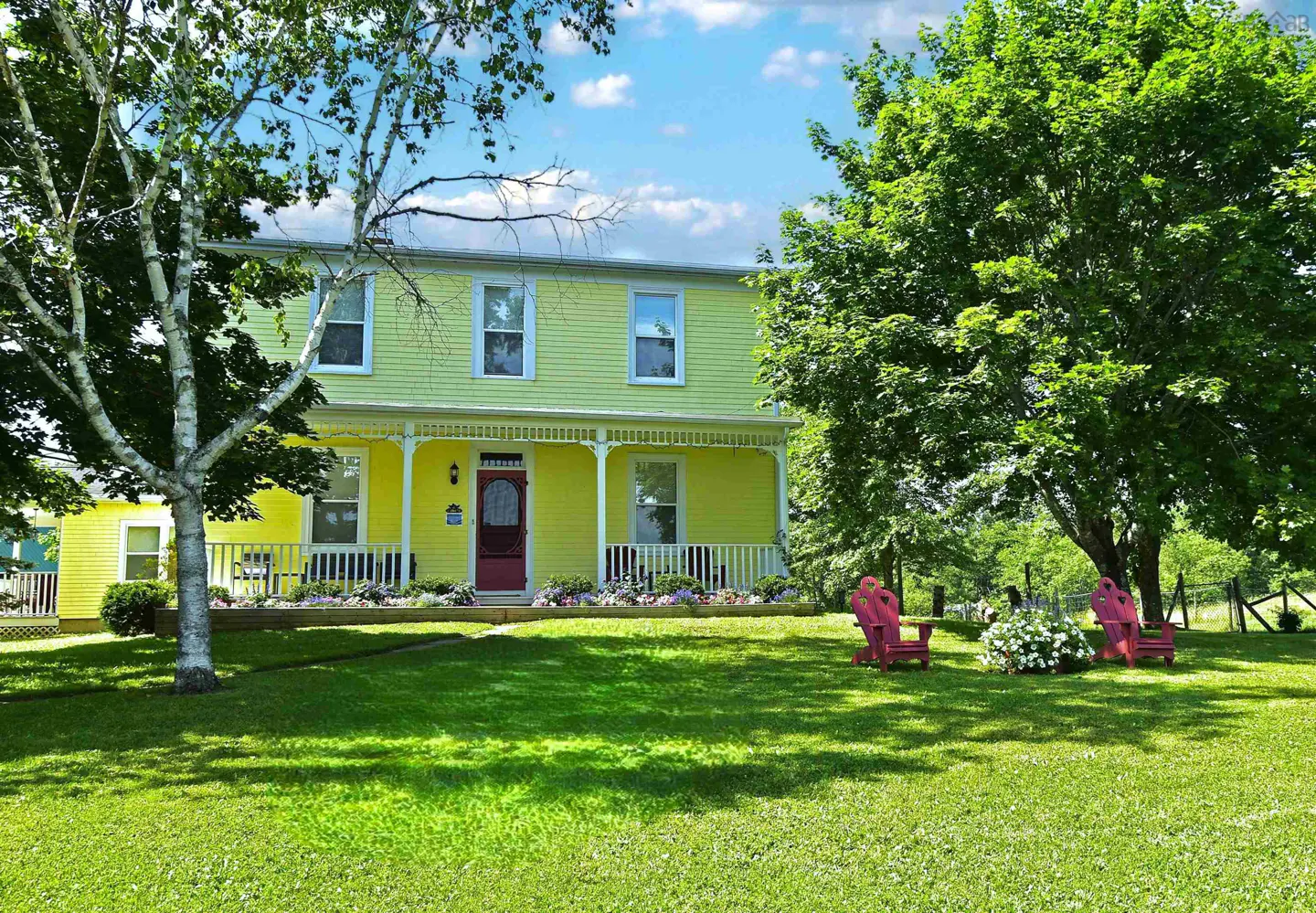 Two-story yellow house with a red door and white porch, surrounded by green lawn and trees. Pink Adirondack chairs sit in the yard.