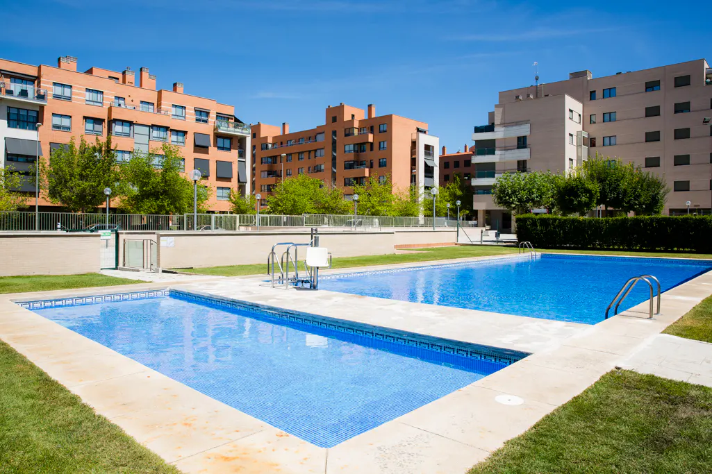 Outdoor swimming pool with blue water, surrounded by green grass and modern apartment buildings under a clear blue sky.