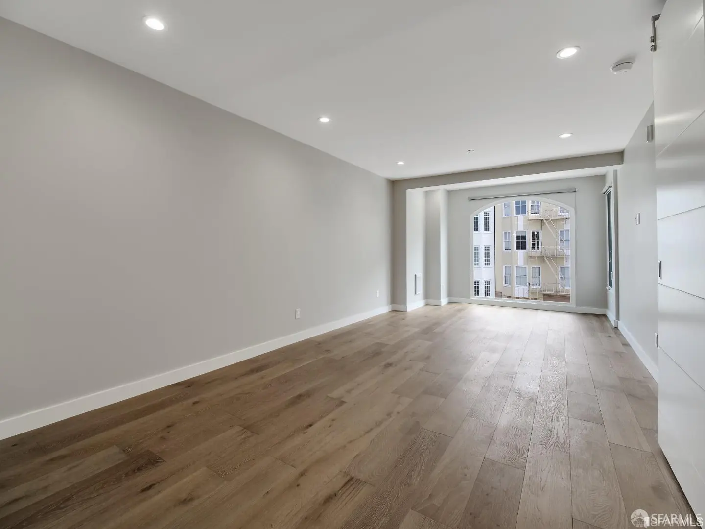 A bright, empty room with light gray walls, wood floors, and a large arched window overlooking a building.