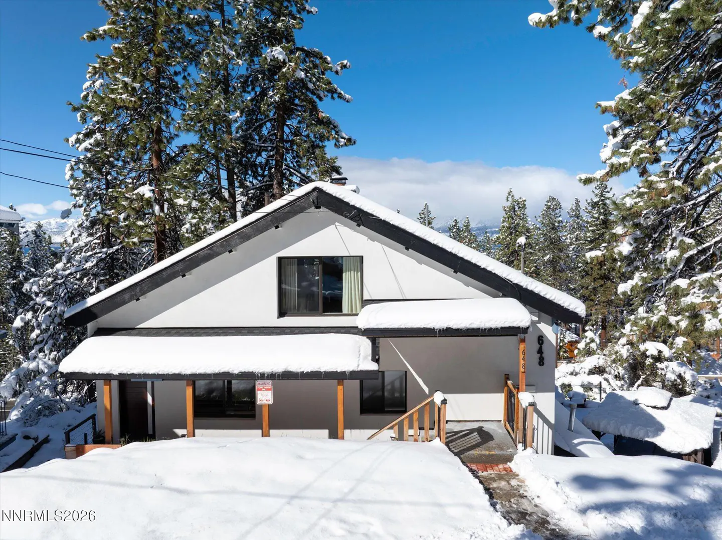 A white house with a black roof is covered in snow, surrounded by snow-covered trees on a sunny day.