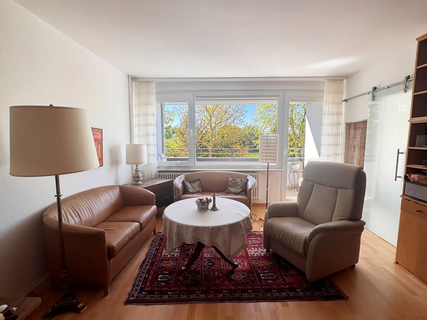 Living room with brown leather sofas, beige armchair, round table with white cloth, and red patterned rug. Large window with trees outside.
