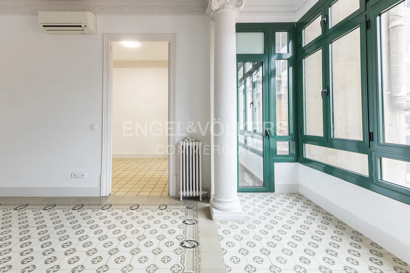 An empty room with white walls, decorative tile floor, green window frames, and a white column. An open doorway leads to another room.