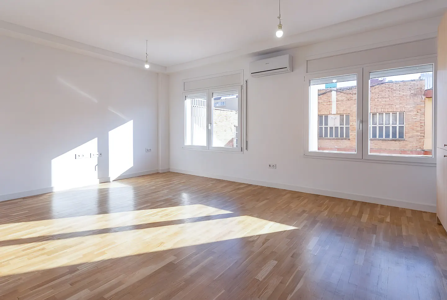 Bright, empty room with hardwood floors, white walls, and two windows showing a brick building.