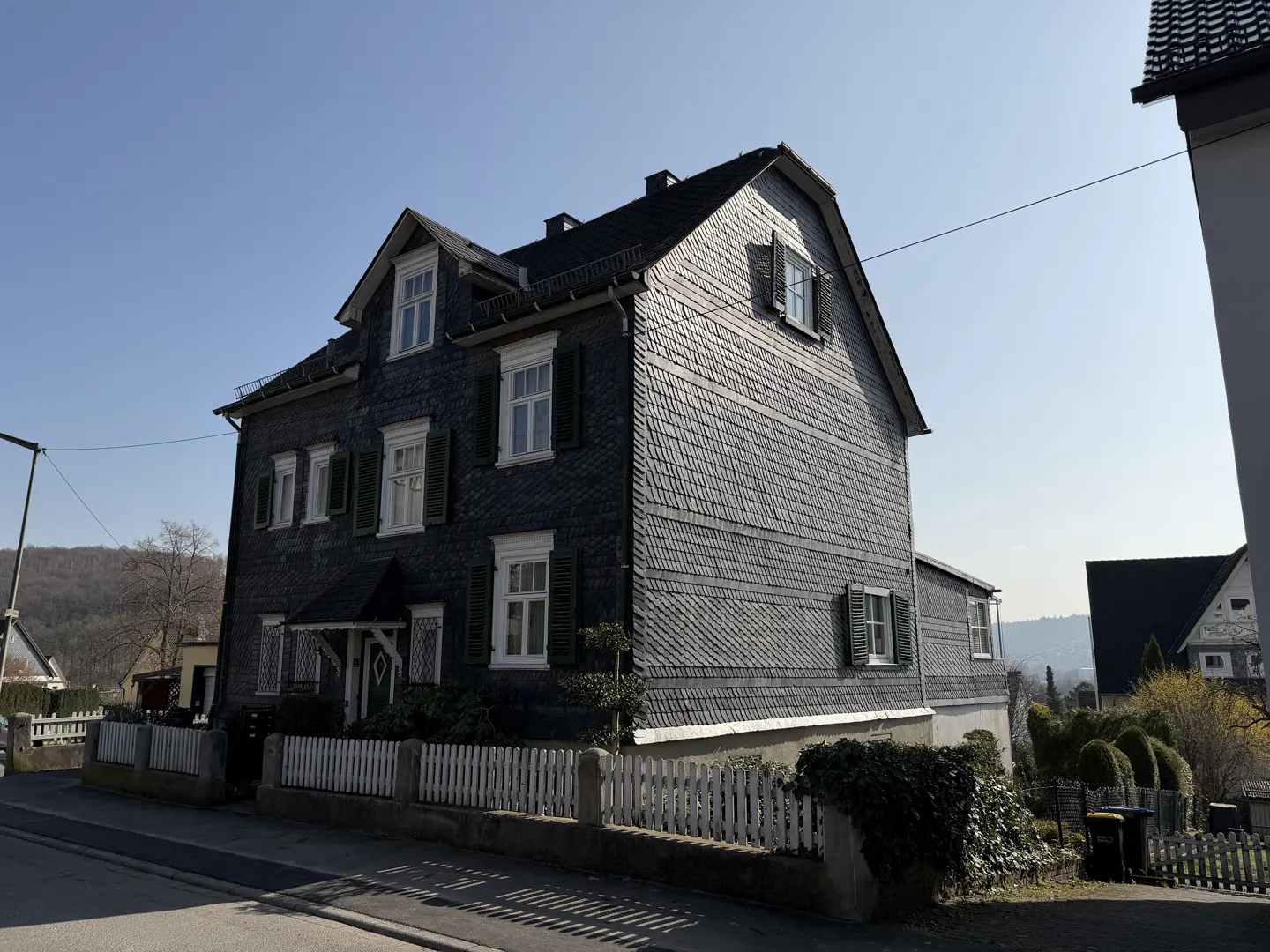 A two-story house with dark gray siding and a white picket fence on a sunny day.