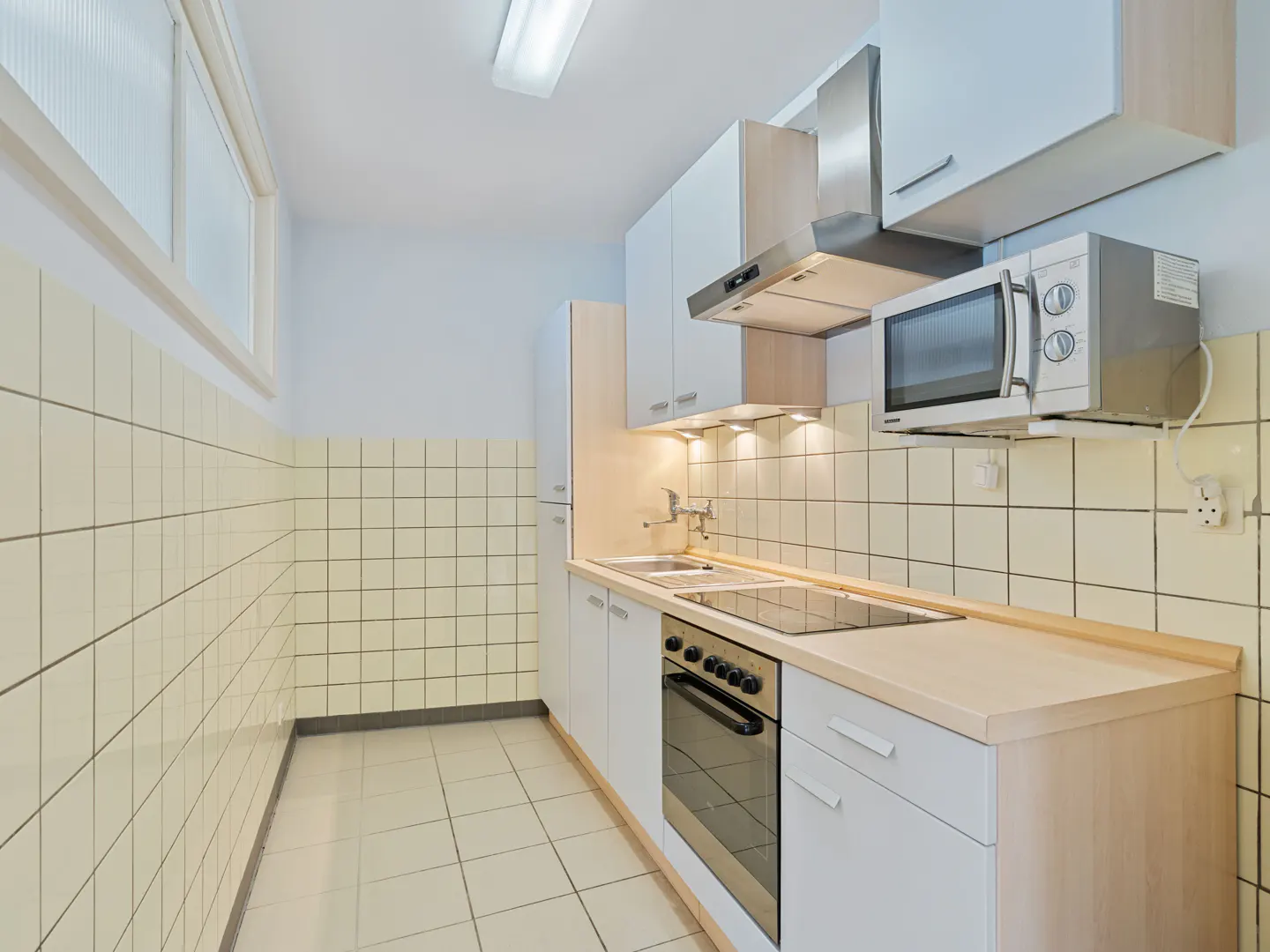 A bright kitchen with white cabinets, beige countertops, and tiled walls and floor. A microwave and range hood are above the stove.