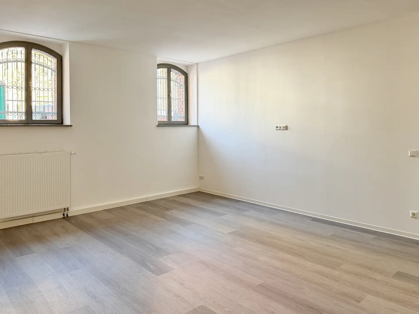 Empty room with gray wood floors, white walls, and two arched windows with decorative iron bars. A white radiator is on the left wall.