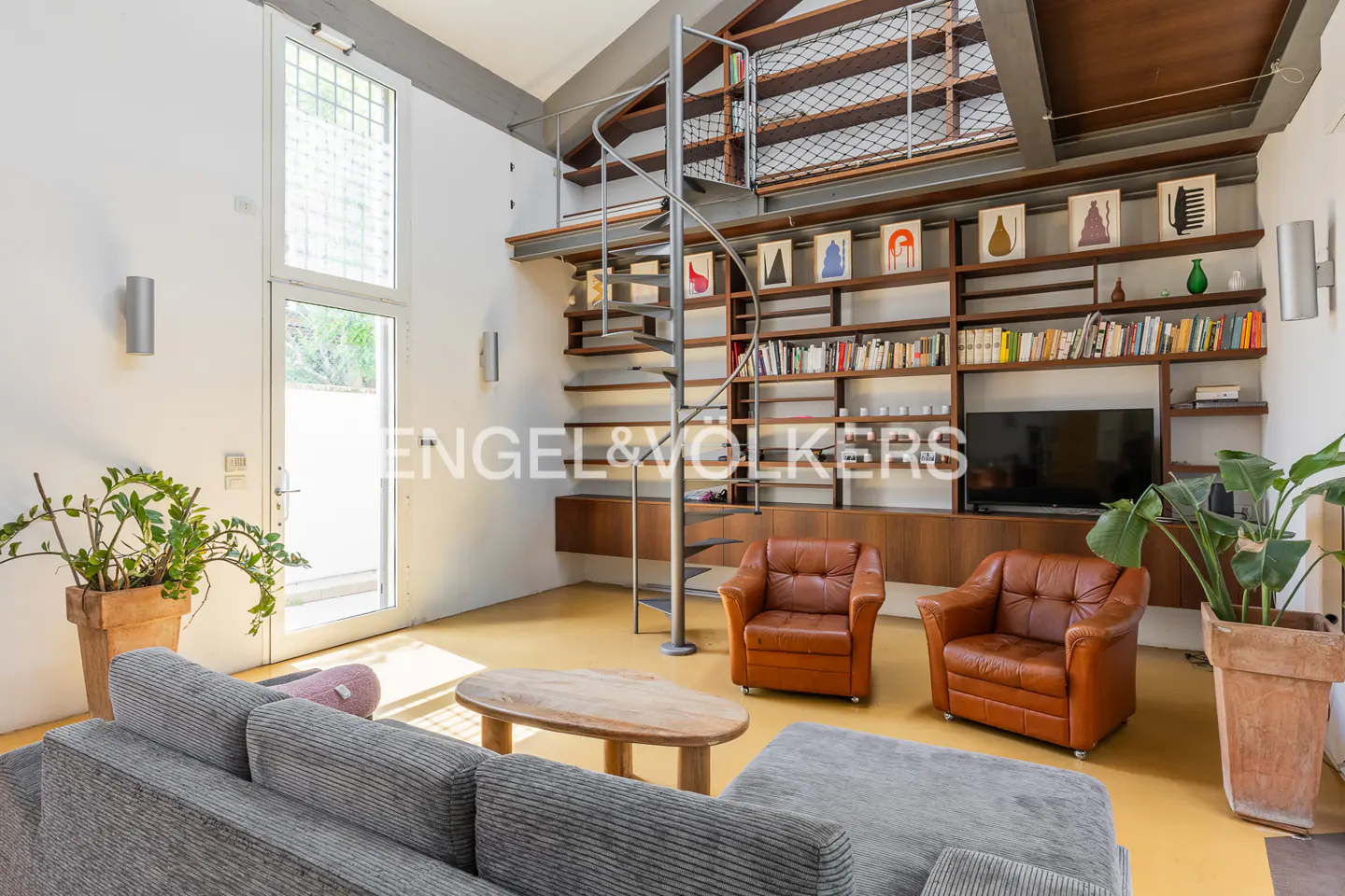 Living room with gray sofa, wood table, and brown leather chairs. A spiral staircase leads to a loft with bookshelves.