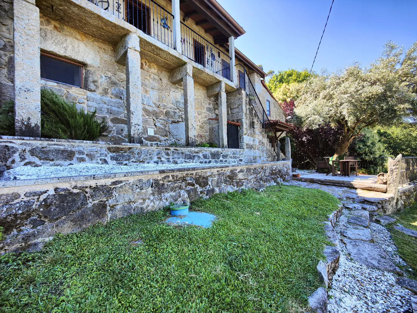 Rustic stone house with a balcony, stone retaining wall, green lawn, and a stone path leading to a patio under trees.