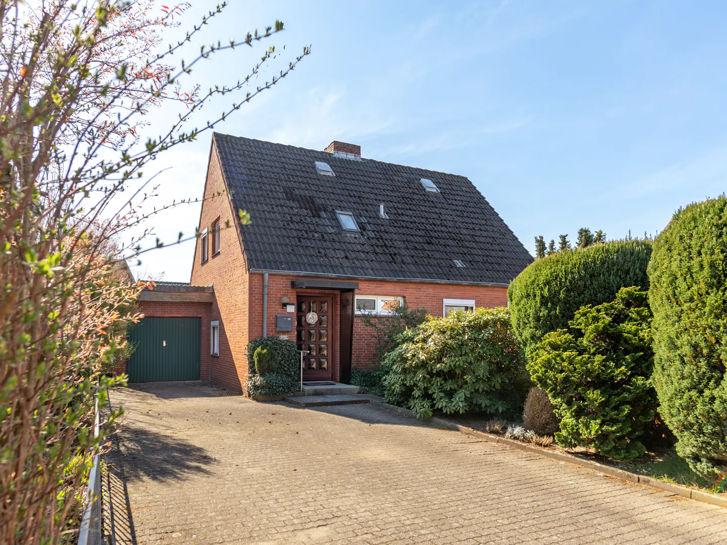 Two-story brick house with a dark roof, green garage door, and paved driveway on a sunny day.
