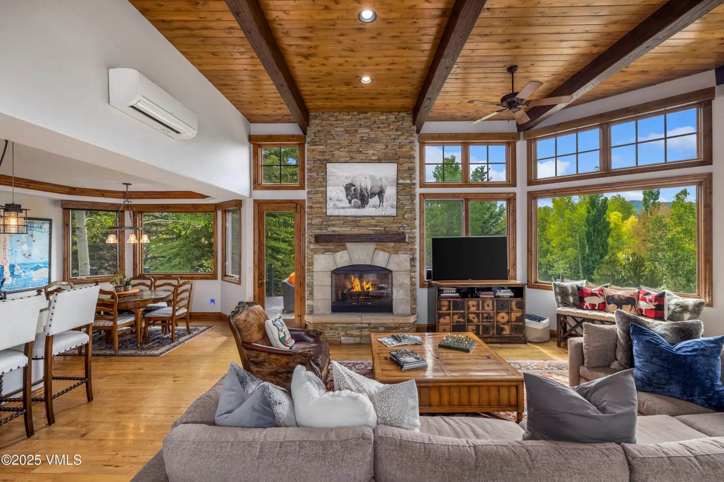 Living room with wood ceiling, stone fireplace, and large windows with green trees outside.
