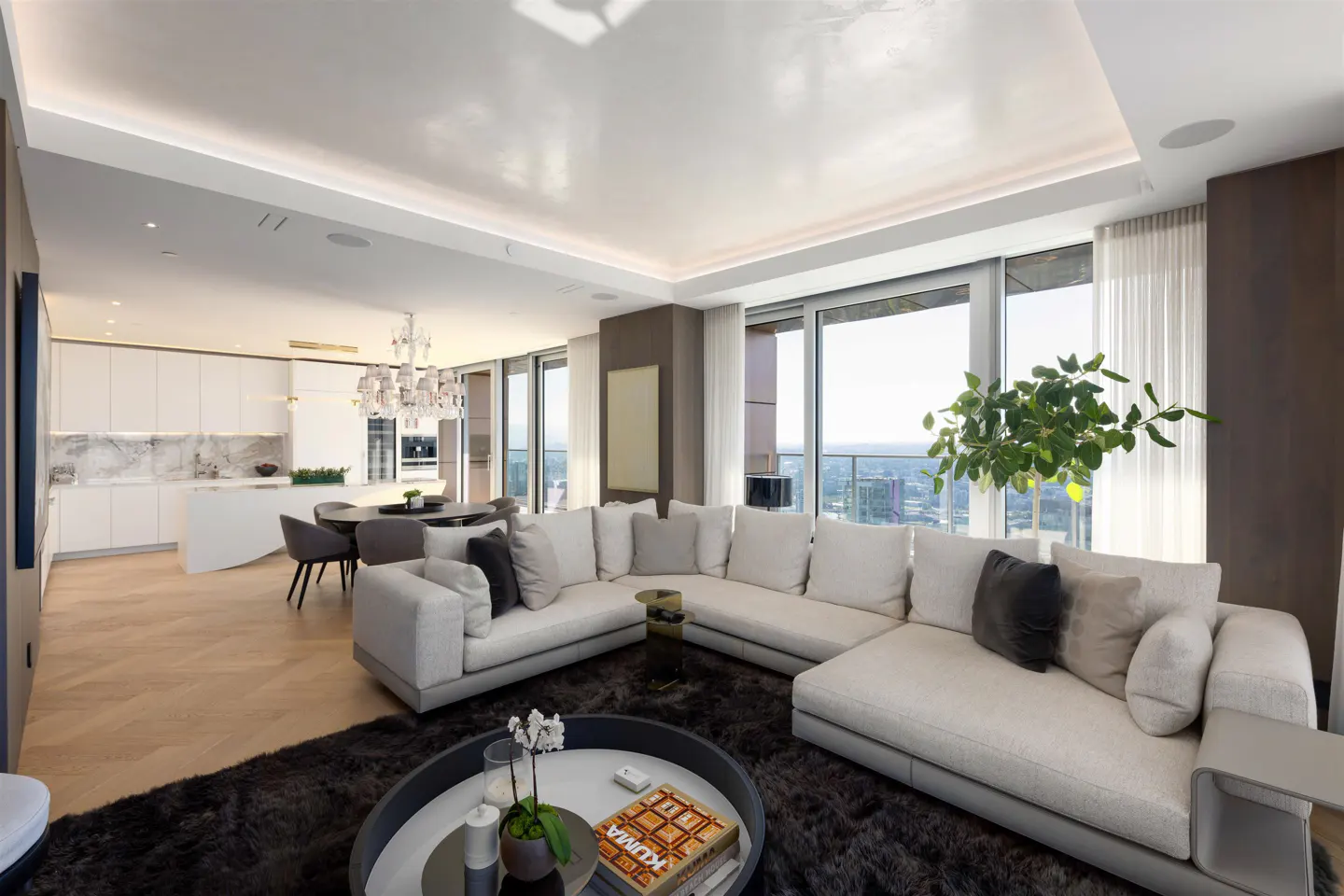 Bright, modern living room with a white sectional sofa, dark rug, and floor-to-ceiling windows overlooking a city view. Open kitchen in the background.