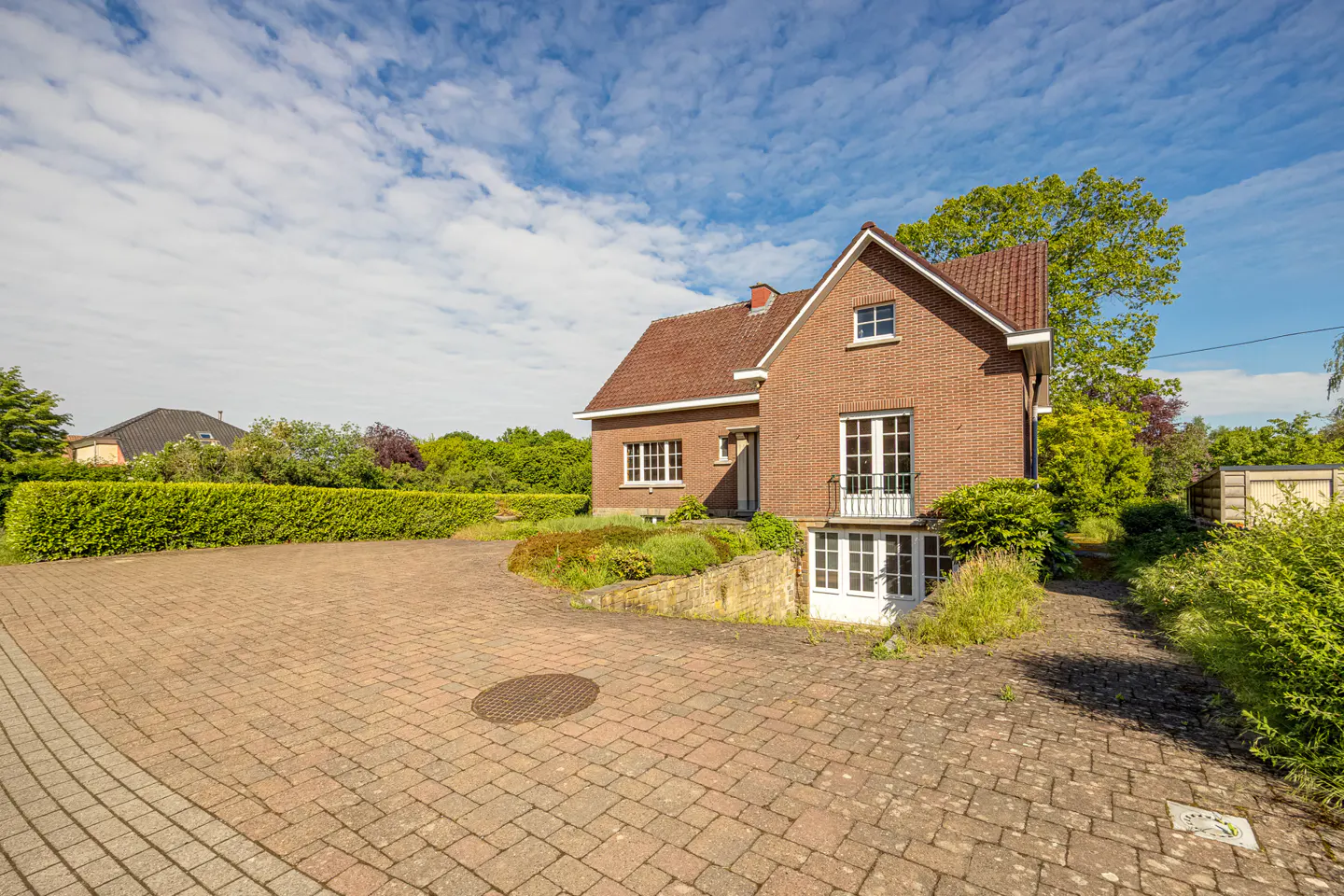 Two-story brick house with a tiled roof, white-framed windows, and a paved driveway under a blue sky with light clouds.