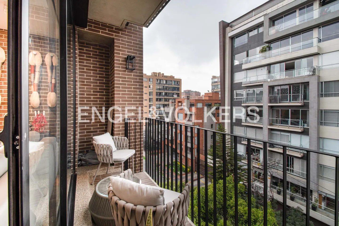 Balcony with brick wall, black railing, and woven chairs with white cushions overlooking city buildings and trees.