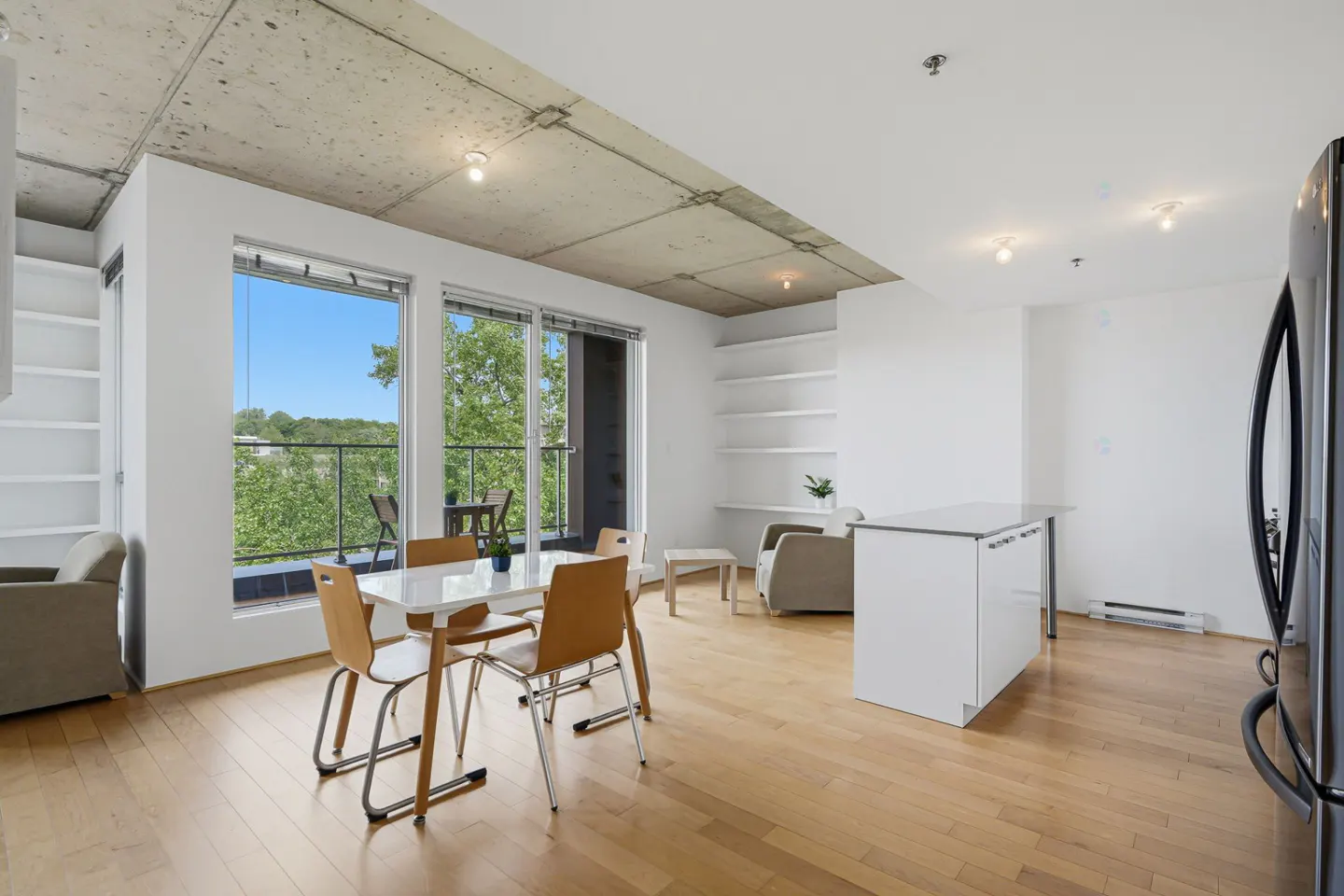 Modern open-concept apartment with concrete ceiling, hardwood floors, dining area, and balcony access with tree views.