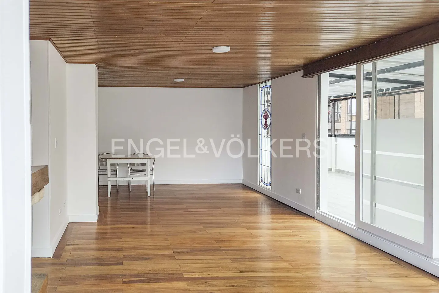 Bright, empty room with wood floors, white walls, and wood ceiling. A stained glass window and sliding glass doors let in light.